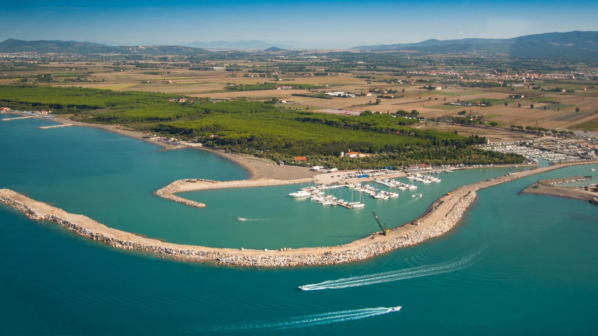 Vista aerea di un porto turistico con barche e frangiflutti, circondato da campagna.