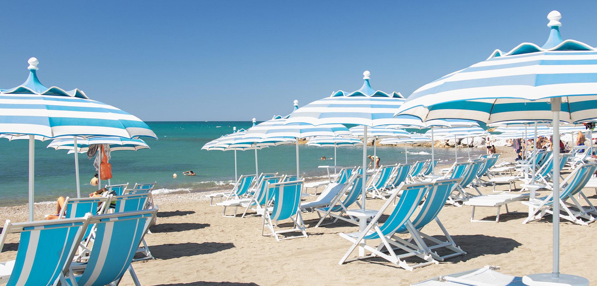 Spiaggia con ombrelloni e lettini azzurri, mare calmo e cielo sereno.