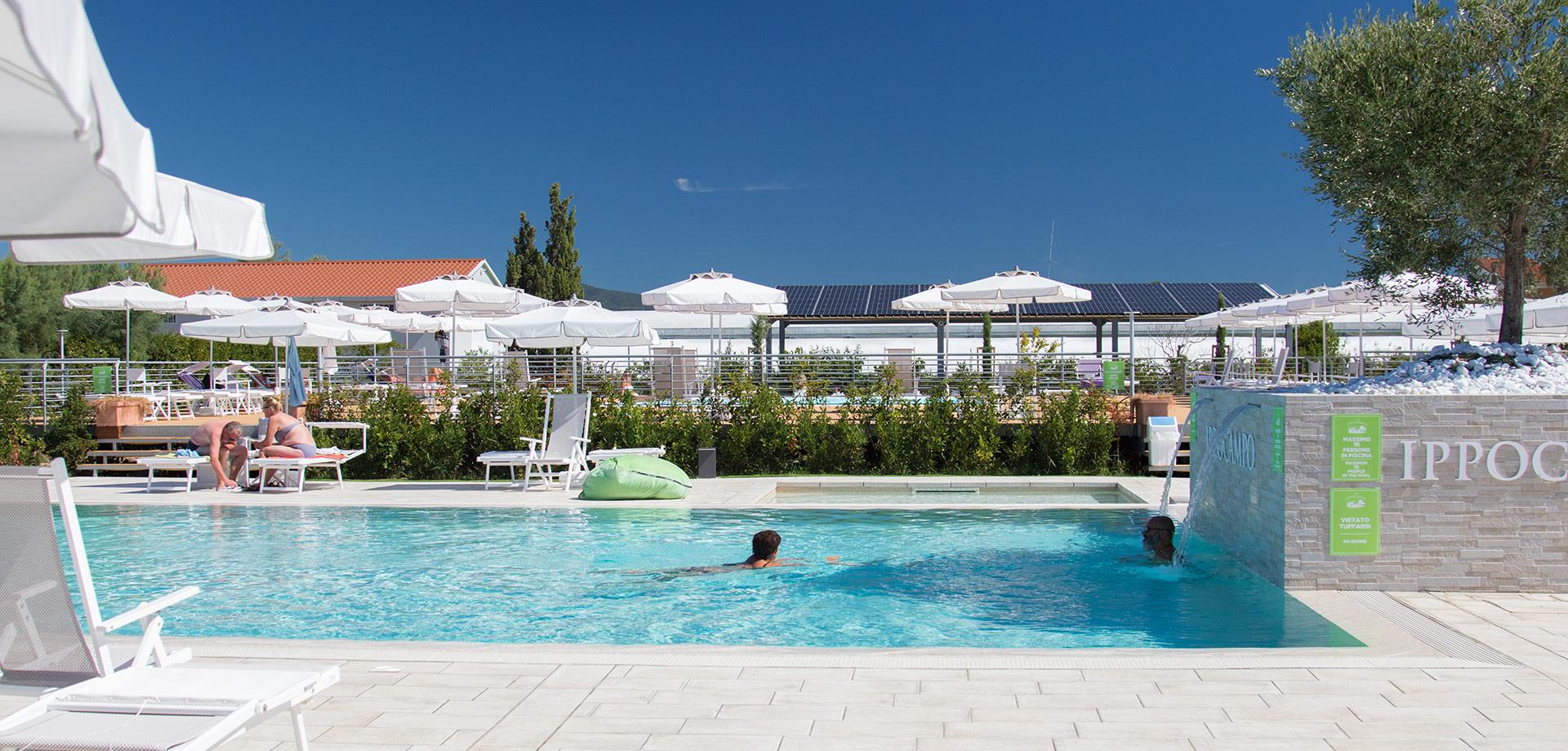 Piscina all'aperto con lettini, ombrelloni e persone che nuotano sotto il sole.