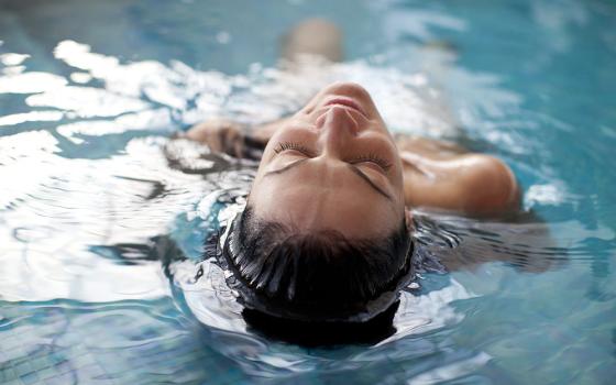 Donna rilassata galleggia in piscina, occhi chiusi, acqua trasparente e calma.