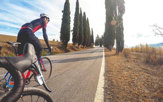 Ciclisti su strada fiancheggiata da cipressi sotto un cielo sereno.