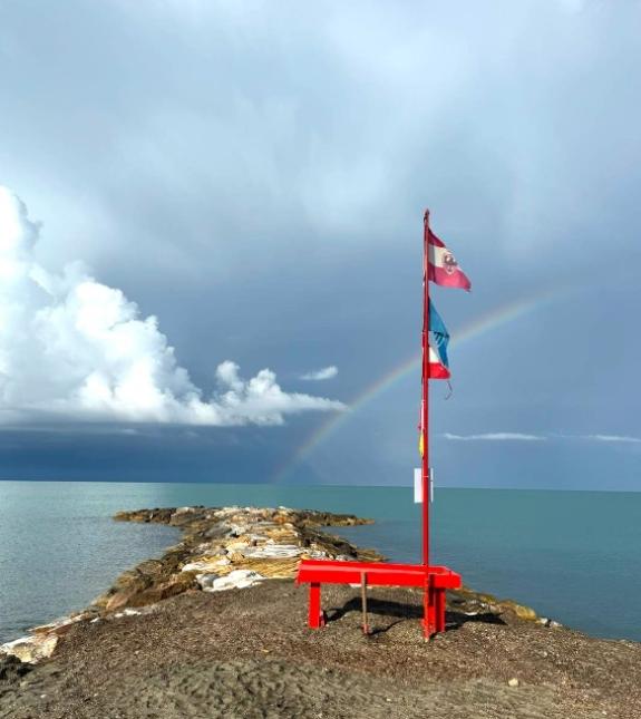 Spiaggia con panchina rossa, bandiere e arcobaleno sopra il mare.