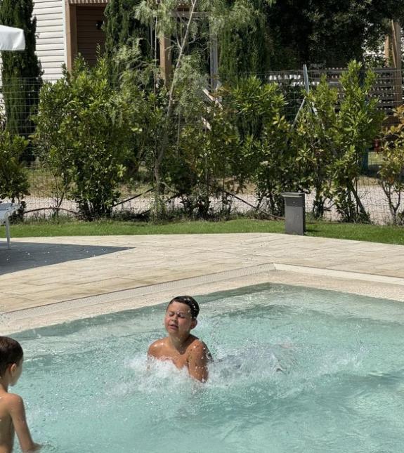 Bambini che giocano in piscina sotto il sole, circondati da vegetazione.