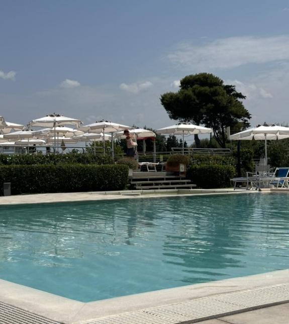 Piscina all'aperto con ombrelloni bianchi e vegetazione circostante, cielo sereno.