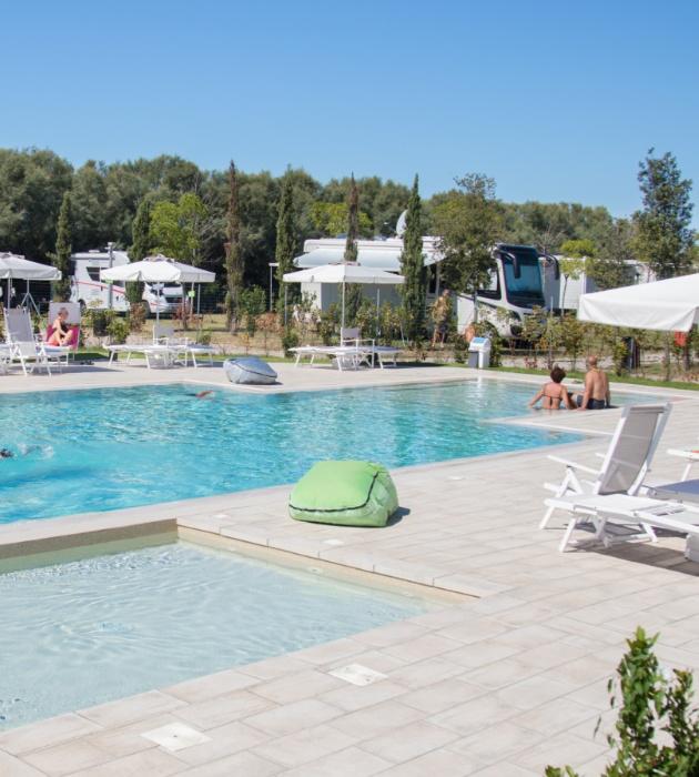 Piscina all'aperto con lettini e ombrelloni, circondata da camper e natura.