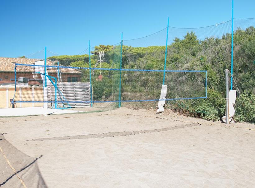 Campo da beach volley con rete e recinzione, circondato da vegetazione.