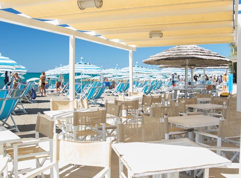 Spiaggia con ombrelloni blu e area ristoro con tavoli e sedie bianche.