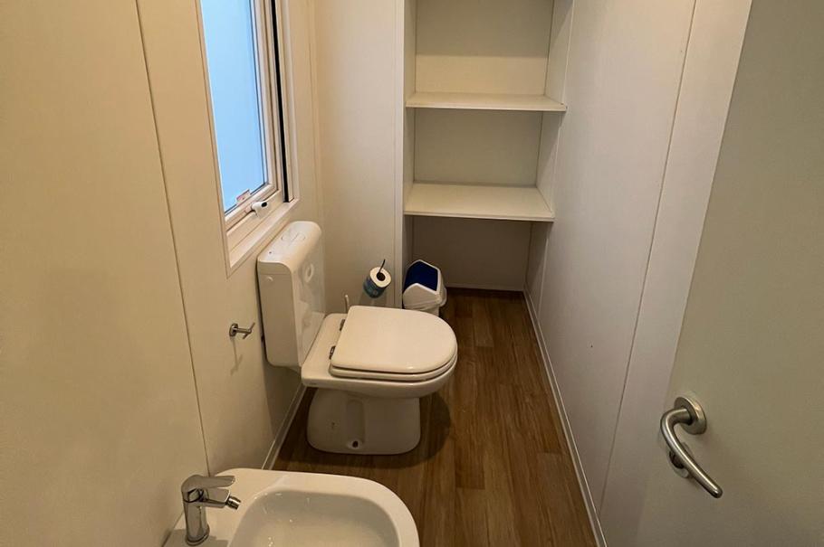 Bagno stretto con wc, bidet, scaffali e finestra, pavimento in legno.