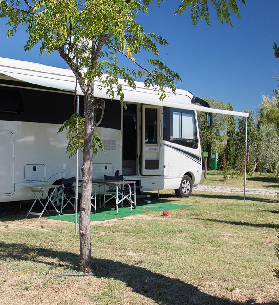 Camper parcheggiato all'ombra, tavolo e sedie all'aperto su prato verde.